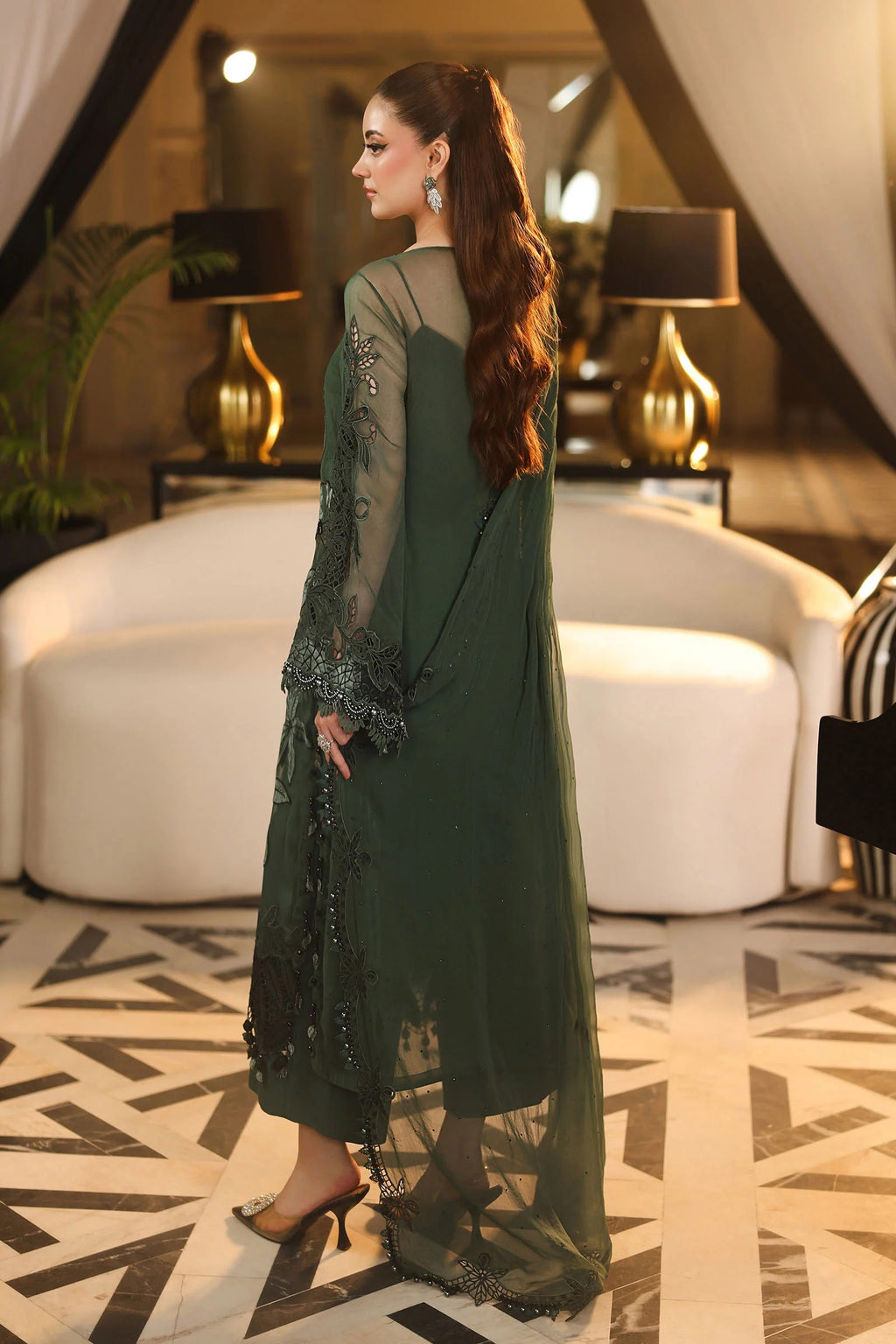 A woman wearing a dark green embroidered traditional dress with sheer sleeves and a long flowing overlay, standing in a modern indoor setting with white curved seating and patterned flooring.