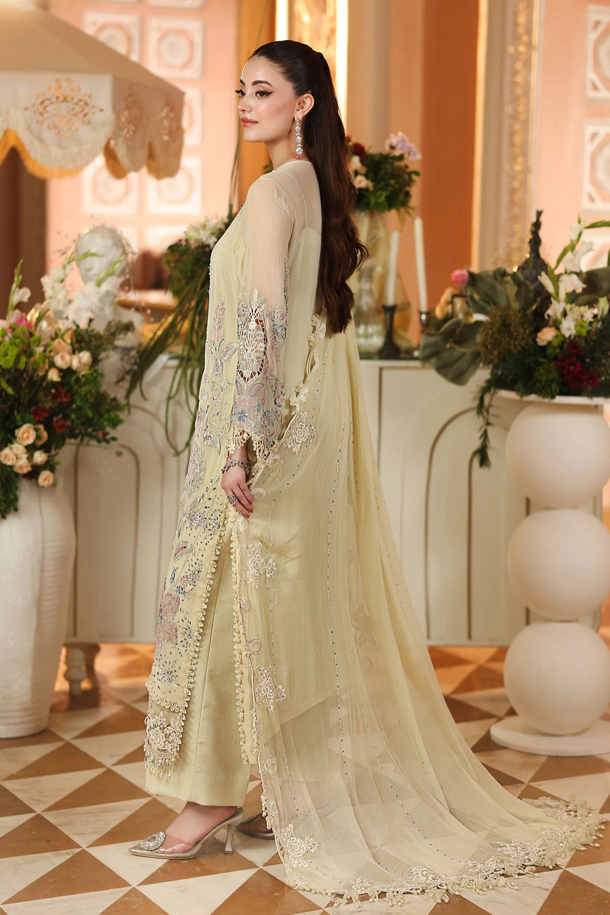 A woman models a traditional South Asian salwar kameez outfit in a warm, decorated indoor setting. The outfit is a light cream/beige color with intricate silver and gold embroidery on the sleeves and hem, paired with a matching sheer dupatta.