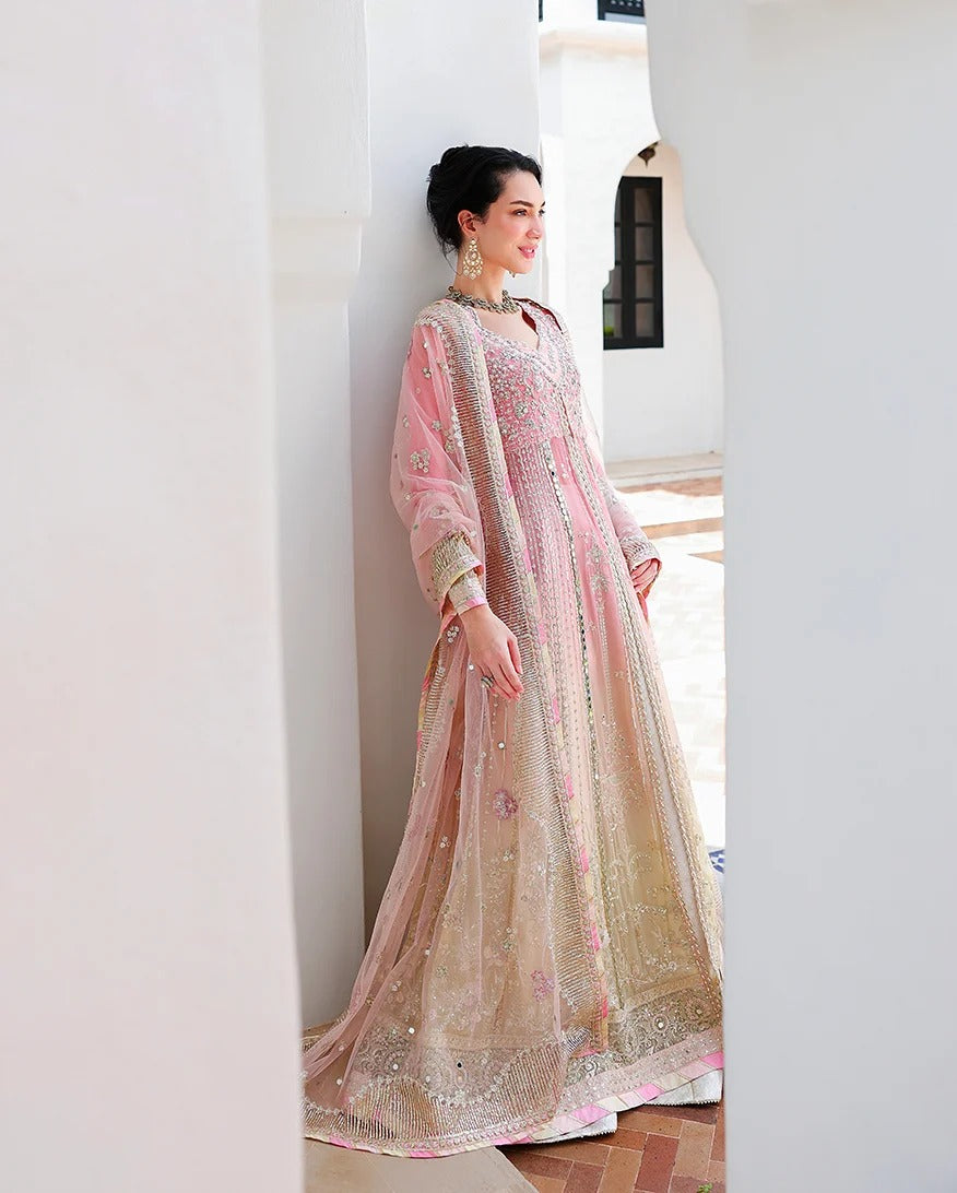 A woman wearing a traditional pink and beige heavily-embroidered Anarkali gown stands in a white architectural corridor.