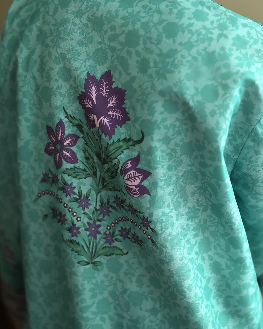 A close-up of a mint green patterned fabric garment, likely a kimono or jacket, featuring a central hand-painted floral motif in purple and green.