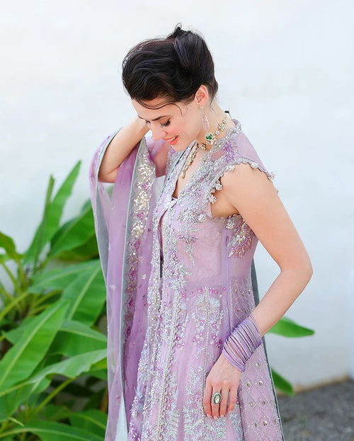 A woman wearing a lavender purple embellished traditional outfit with silver embroidery, a matching dupatta, and bangles, standing outdoors against a white wall with green plants on the left.