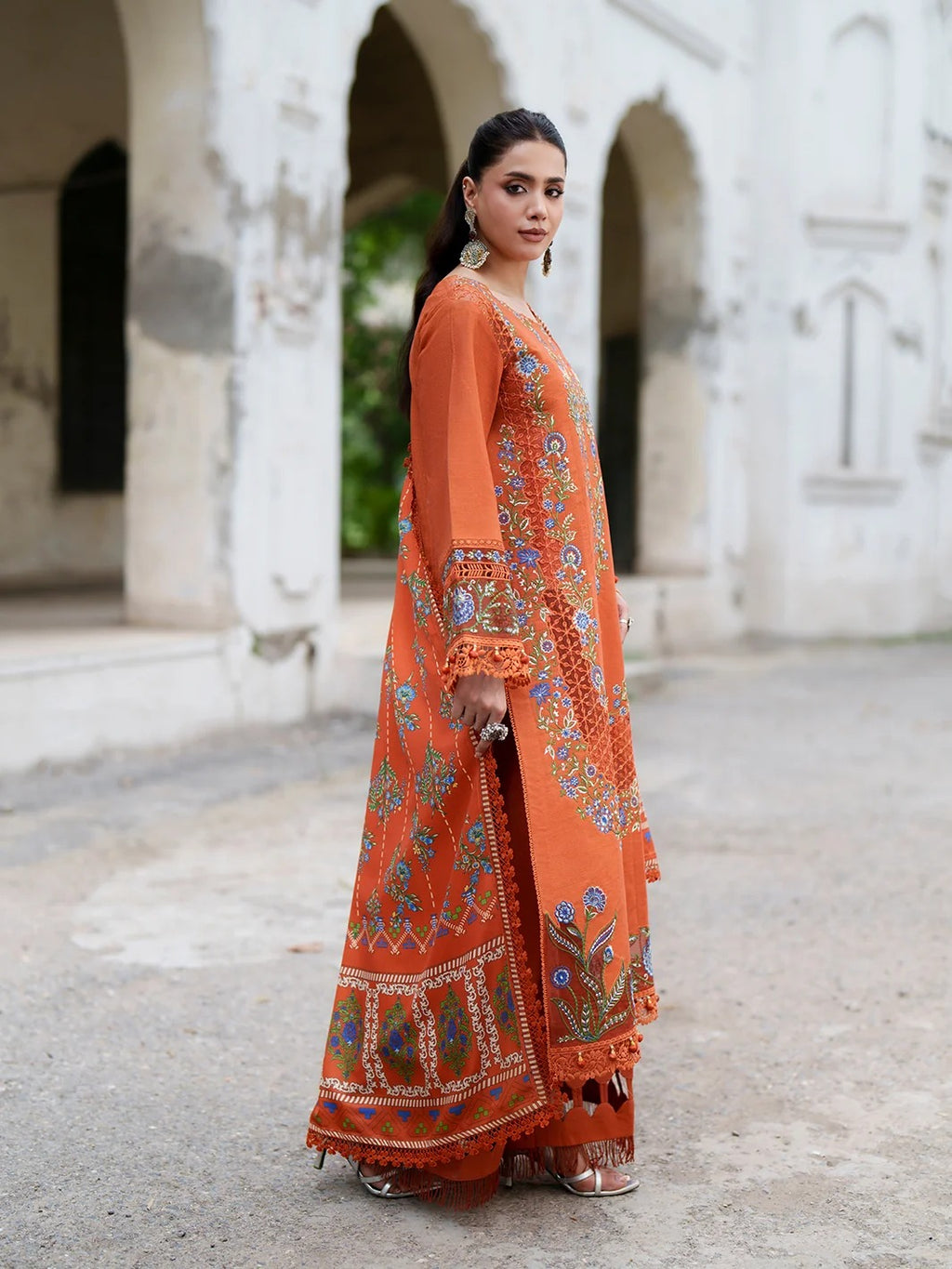 A woman models a traditional Indian outfit consisting of an orange anarkali kurta and matching palazzo pants, both heavily decorated with colorful floral embroidery and mirror work, set against a blurred background of a white archway building.