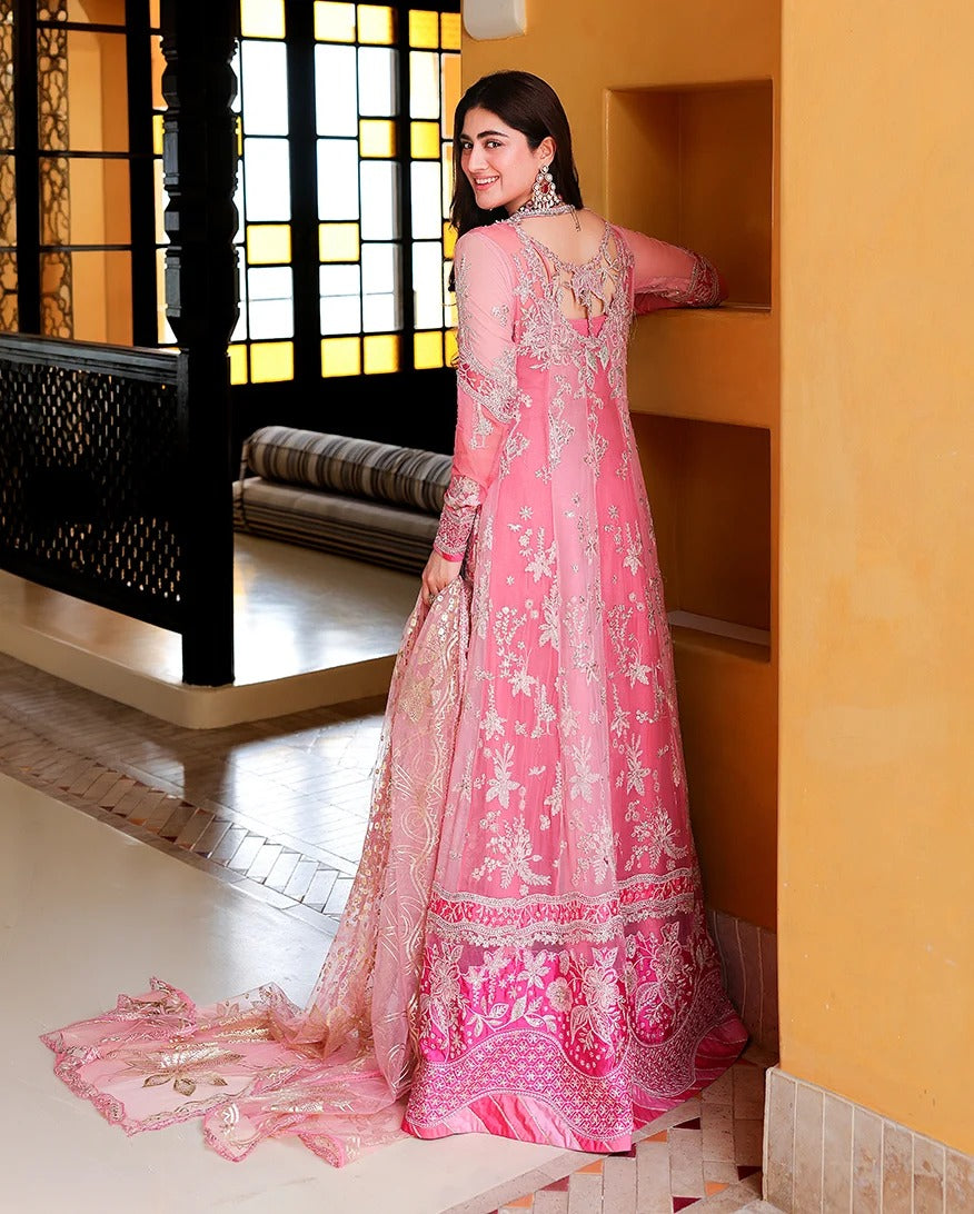 A woman wearing a pink embroidered traditional gown with a long train, standing in an indoor architectural setting with warm yellow walls and window light.