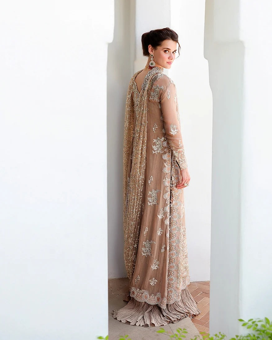 A woman wearing a beige embroidered traditional gown with a matching dupatta, photographed from the back in a bright white architectural setting.