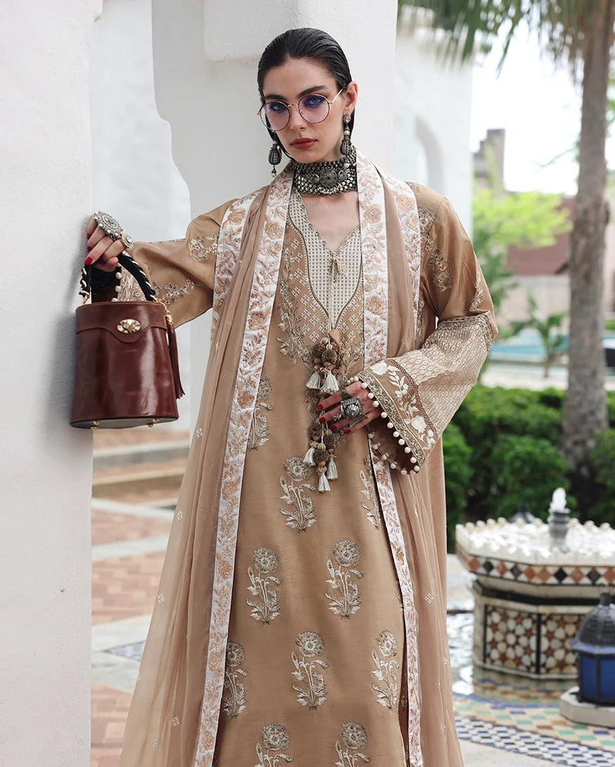 A woman models a traditional beige embroidered outfit with a matching sheer outer layer, accessorized with a brown leather handbag, statement earrings, and a pearl choker in an outdoor courtyard setting.