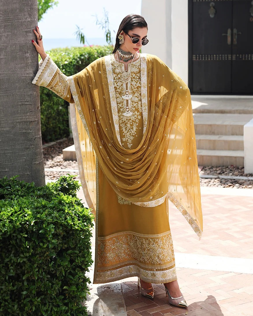 A woman models a traditional Indian salwar kameez suit in mustard yellow with intricate gold embroidery and a matching sheer dupatta, set against an outdoor residential background.