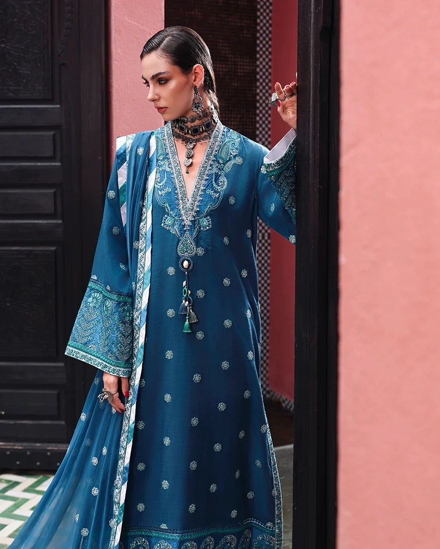 A woman models a traditional blue three-piece salwar kameez suit with intricate silver embroidery, a matching dupatta, and a long necklace, standing in a doorway with a pink wall background.