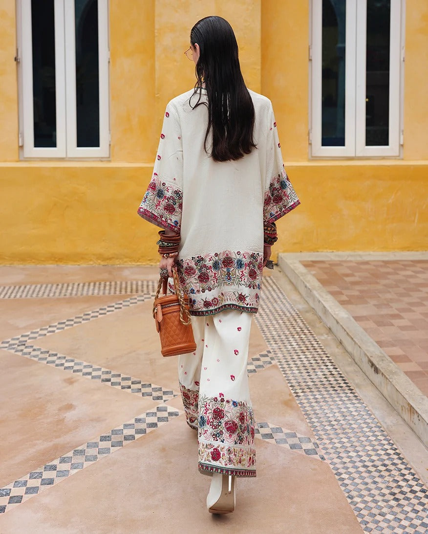A woman viewed from behind walking outdoors in a cream-colored embroidered tunic and matching wide-leg pants, carrying a brown woven handbag against a yellow building wall.