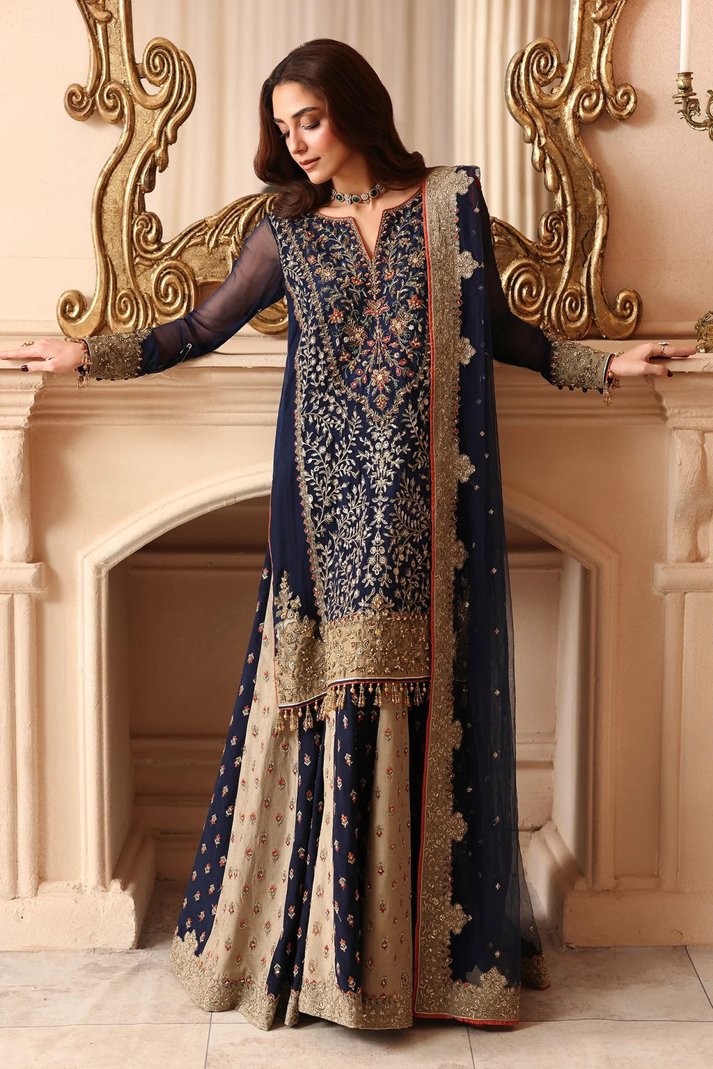 A woman models a traditional Indian navy blue and beige embroidered outfit with a matching dupatta, posed in front of an ornate beige fireplace with gold detailing.