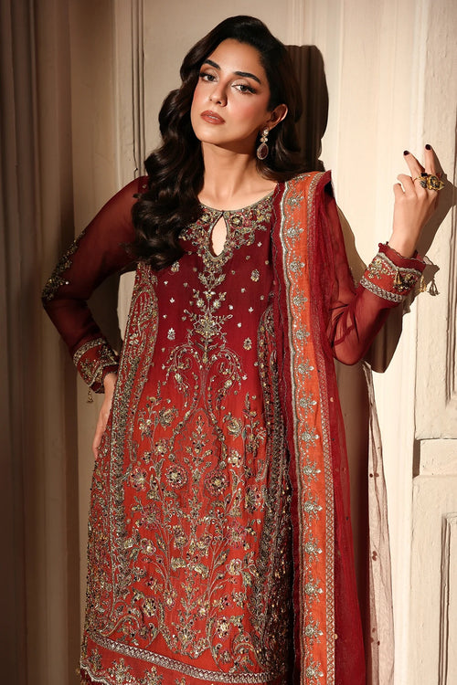 A woman models a traditional red and gold embroidered Indian salwar kameez suit with a matching dupatta, posed against a neutral beige background.