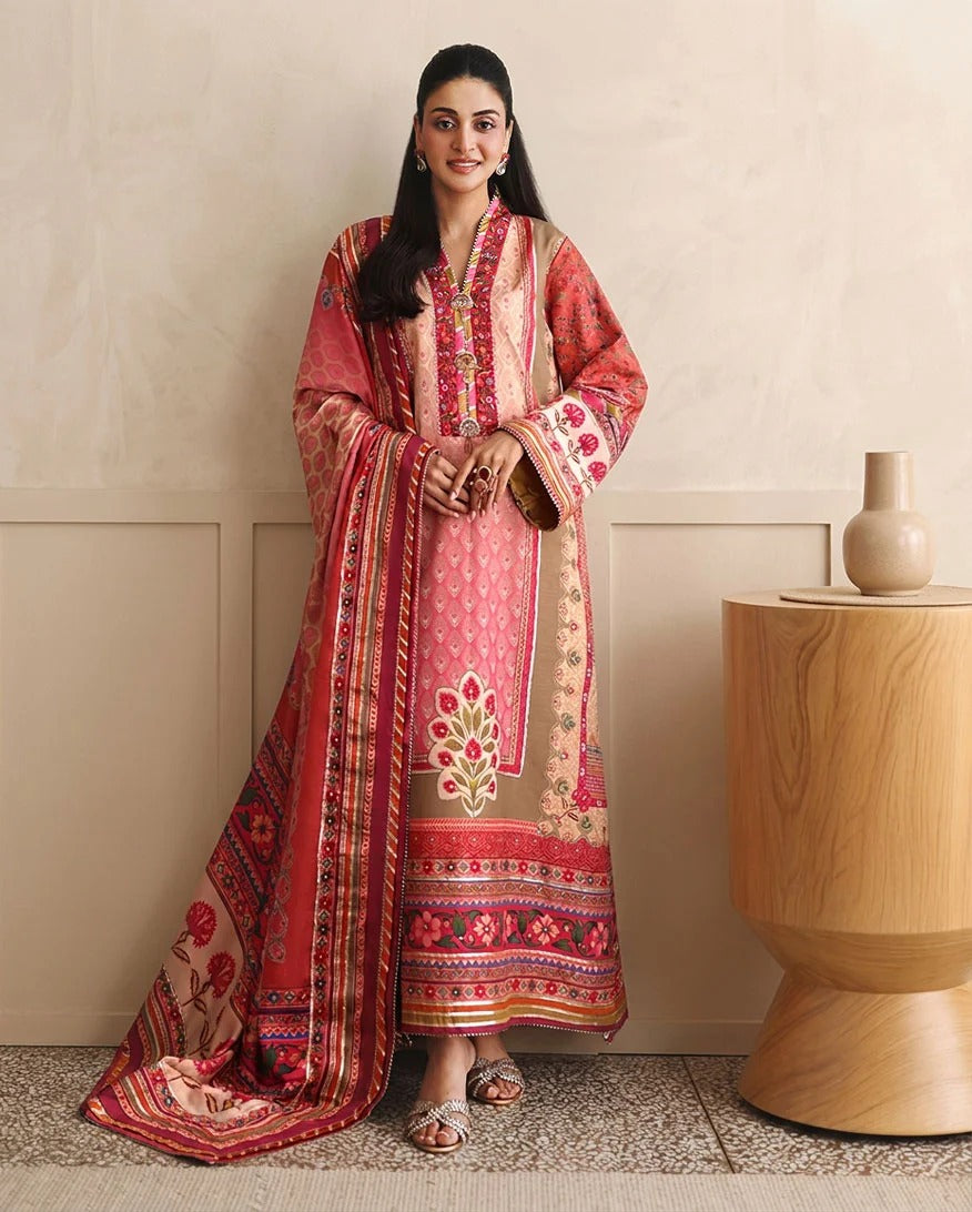 A woman models a traditional South Asian salwar kameez suit in a warm-toned indoor setting. The outfit features a pink and beige kurta with intricate floral embroidery and a matching dupatta draped over the floor, styled with a wooden side table and neutral wall background.