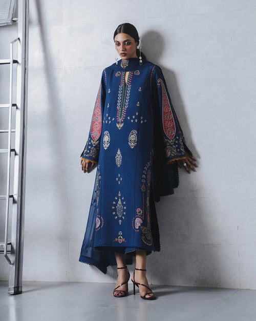 A woman models a long blue embroidered tunic with wide sleeves and intricate red, gold, and white patterns, standing against a light gray concrete wall.