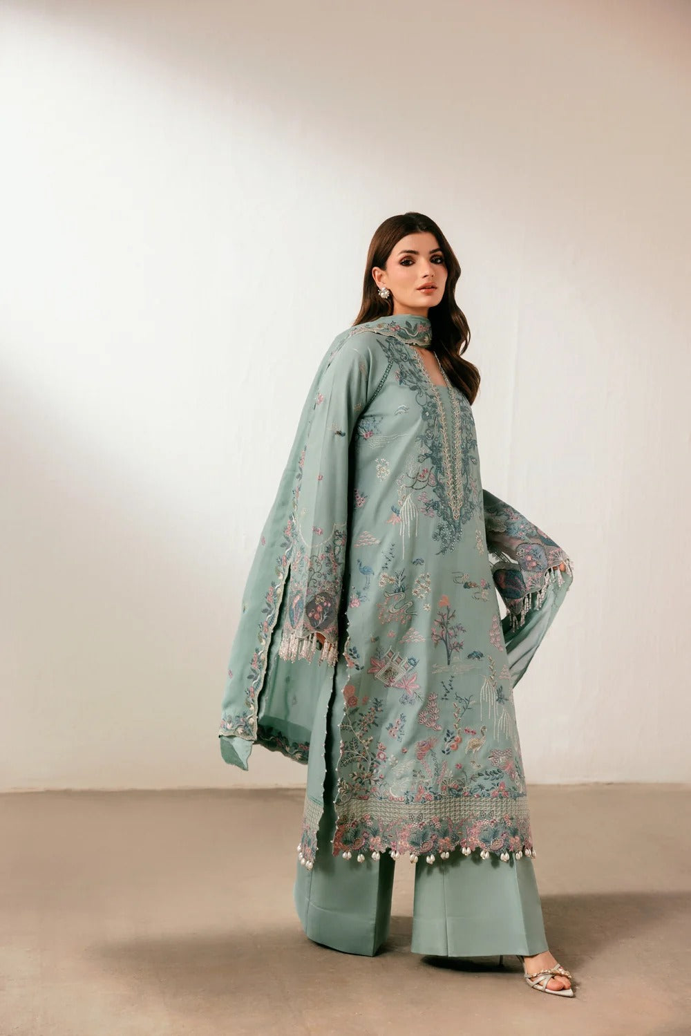 A woman models a three-piece mint green ethnic suit with floral embroidery and tassel borders, consisting of a long kurta, wide-leg trousers, and a matching dupatta, set against a plain beige studio background.