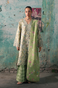A woman models a traditional South Asian outfit consisting of a light sage green kurta with floral embroidery, matching wide-leg trousers, and a coordinating light green dupatta, set against a rustic, textured teal wall.