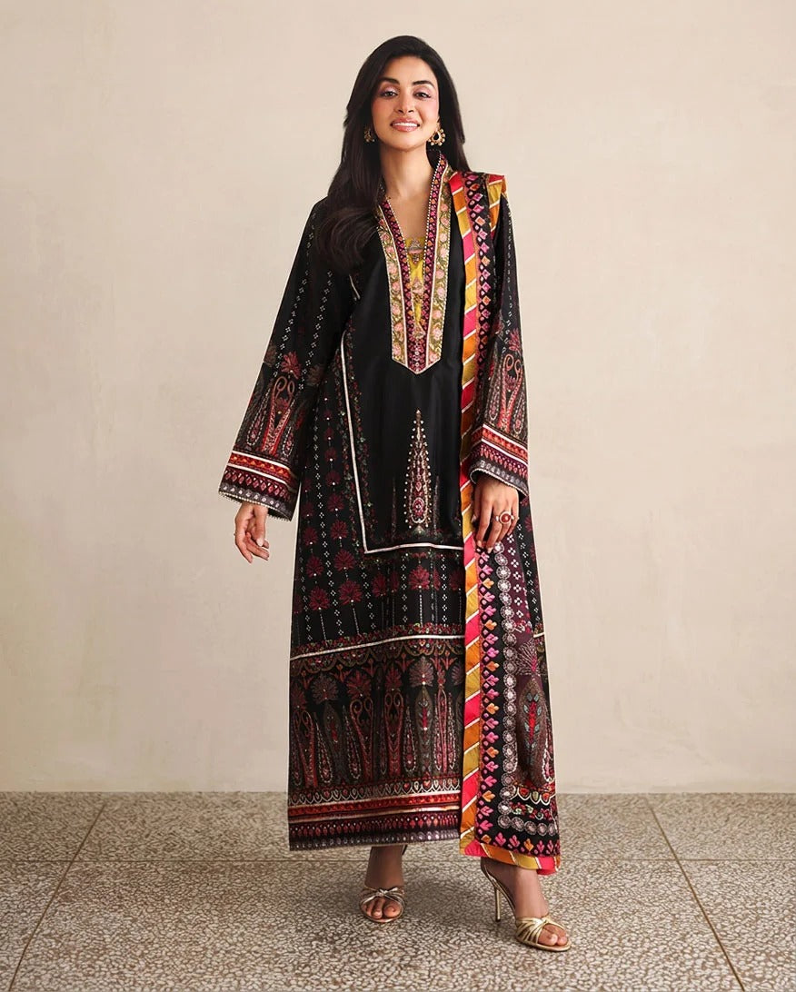 A woman models a traditional black kurta set with vibrant red, yellow, and gold patterns, paired with a matching printed dupatta, set against a neutral beige wall and tiled floor.