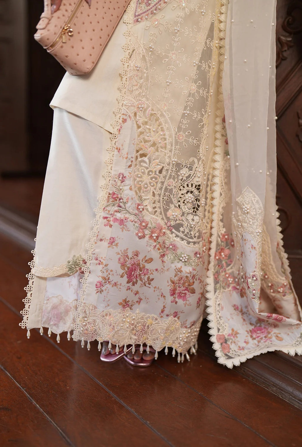 A woman with long dark hair wearing a cream-colored embellished traditional outfit with floral embroidery and pearl necklaces, posing in a softly lit indoor setting.