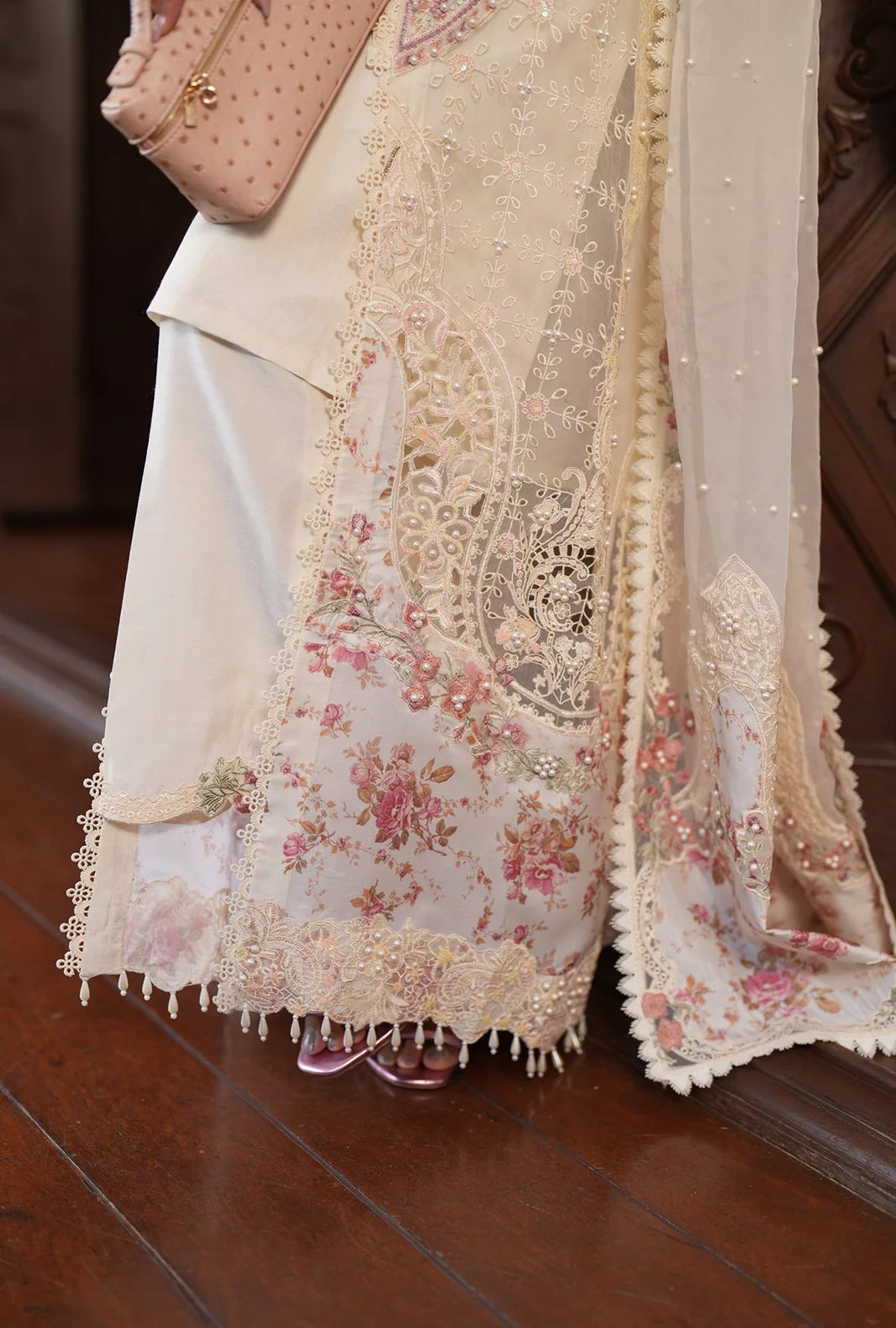 A woman with long dark hair wearing a cream-colored embellished traditional outfit with floral embroidery and pearl necklaces, posing in a softly lit indoor setting.