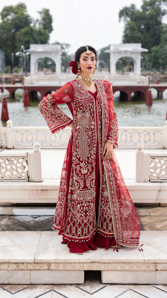 Woman in a red embroidered traditional outfit standing in front of a scenic background with water and architecture.