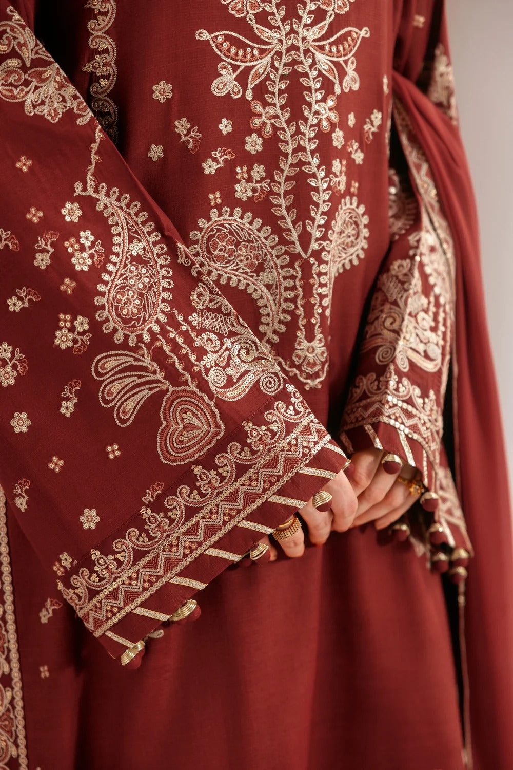 A close-up of a maroon traditional embroidered garment with intricate cream paisley and floral embroidery, featuring wide sleeves and a draped dupatta, with hands holding the hem.
