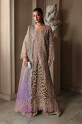 A woman models a heavily embellished beige and lavender traditional gown with intricate floral embroidery and beadwork, holding a small silver clutch in an elegant indoor setting with a classical bust statue in the background.