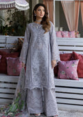 A woman models a light lavender-gray three-piece traditional outfit with intricate embroidery and lace, paired with matching wide-leg trousers and a sheer dupatta with floral prints, posed in front of a white wooden bench with pink cushions.