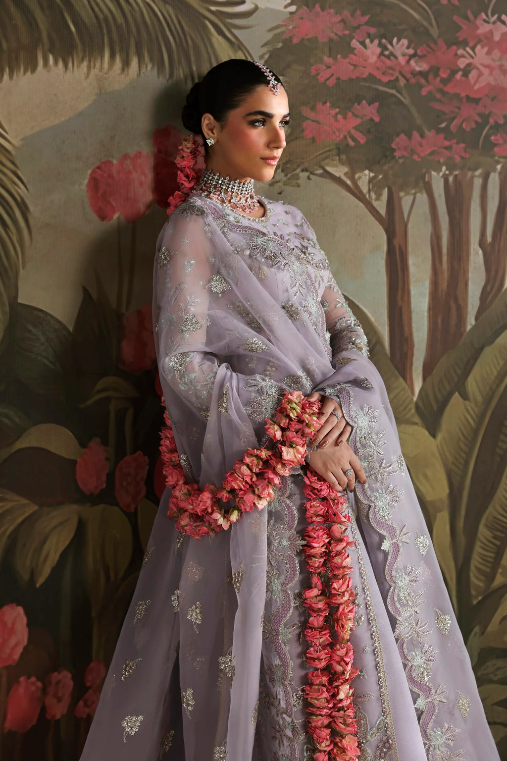 A woman models a lavender purple embroidered traditional outfit with pink floral garlands, standing against a painted tropical foliage backdrop.