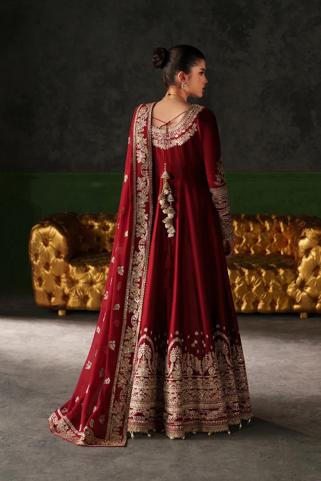 A woman stands with her back to the camera wearing an ornate deep red traditional gown with extensive gold embroidery and a matching embroidered dupatta. She is posed in front of a dark textured wall and a gold tufted sofa.