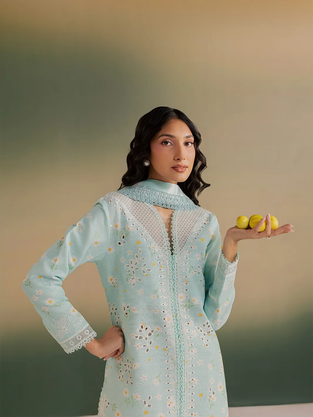 A woman models a light mint green embroidered kurta with a matching dupatta and a small white embroidered border. She is holding three yellow lemons in her hand against a soft, warm-toned gradient background.