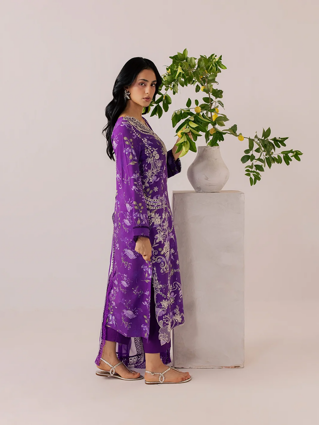 A woman modeling a purple embroidered kurta with matching pants and flat sandals, standing next to a white pedestal with a green plant in a studio setting.