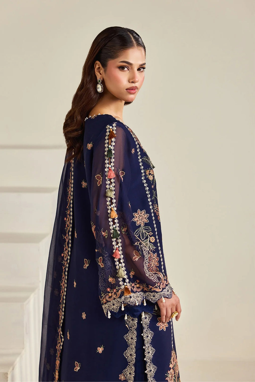 A woman modeling a navy blue embroidered traditional outfit with sheer long sleeves and intricate floral embroidery in gold, orange, and white, set against a neutral beige background.