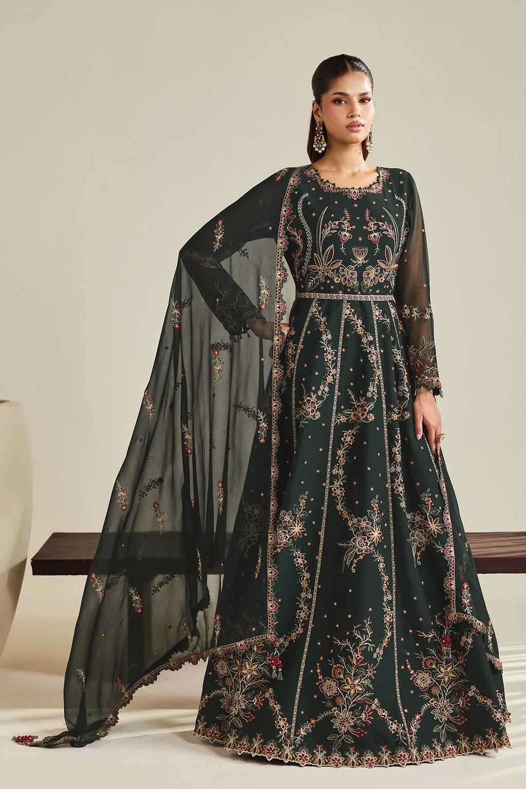 A model wears a dark forest green anarkali gown with intricate gold, beige, and red floral embroidery, paired with a matching sheer black dupatta, set against a neutral studio background.