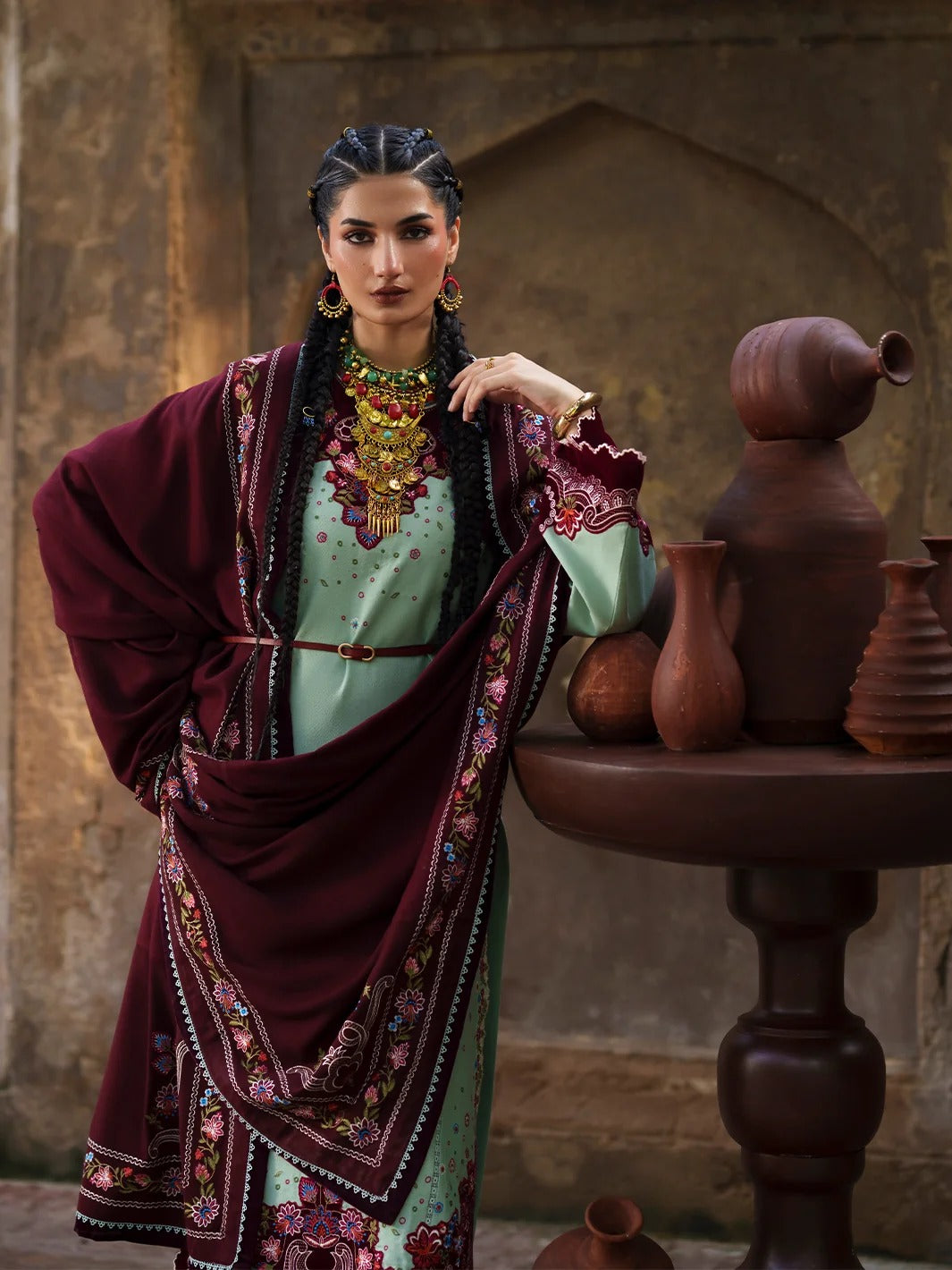 A woman models a traditional South Asian outfit featuring a mint green embroidered kurta with a deep maroon shawl draped over her shoulders, set against a rustic stone wall with clay pottery on a table.
