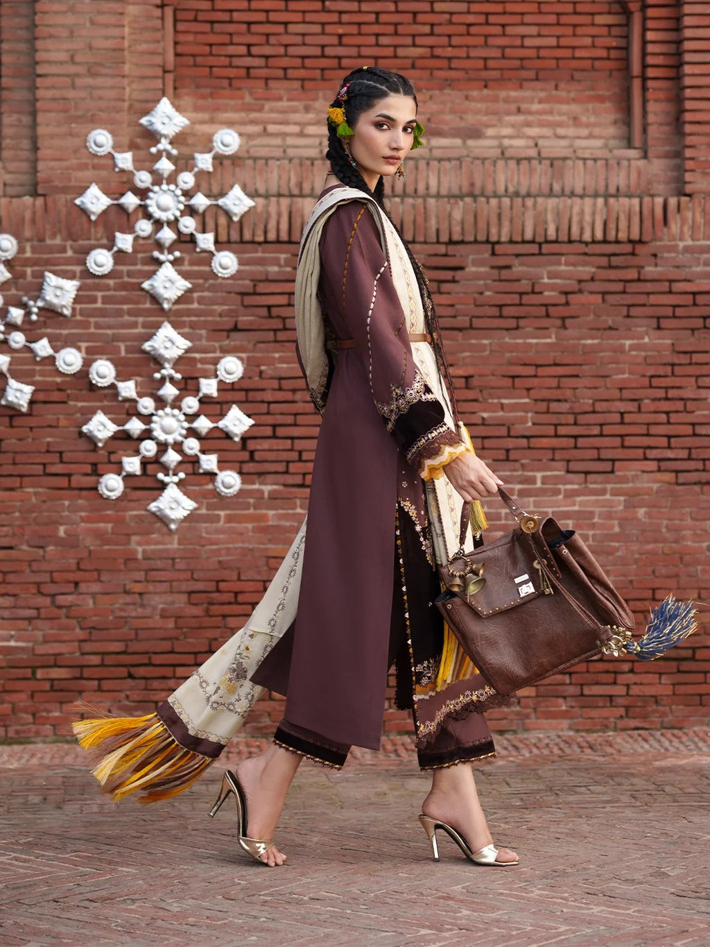 A woman models a traditional South Asian outfit featuring a long brown tunic with beige and gold patterns, a matching brown dupatta, and wide-leg trousers with fringe detailing. She is walking in front of a red brick wall decorated with a white metal snowflake ornament, carrying a brown leather handbag.
