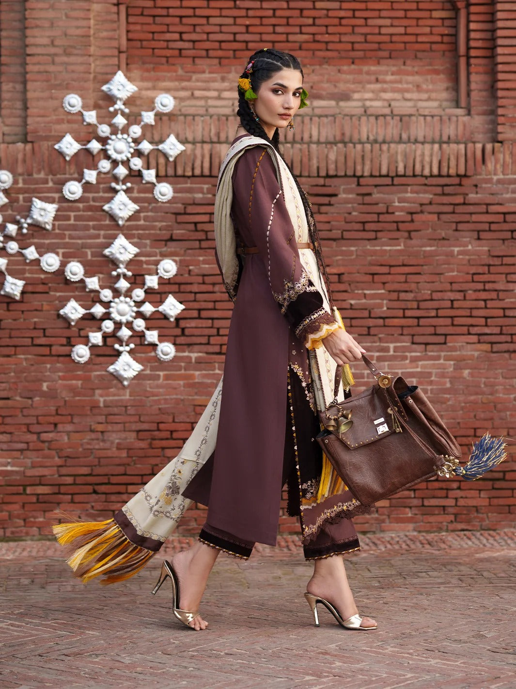 A woman models a traditional South Asian outfit featuring a long brown tunic with beige and gold patterns, a matching brown dupatta, and wide-leg trousers with fringe detailing. She is walking in front of a red brick wall decorated with a white metal snowflake ornament, carrying a brown leather handbag.