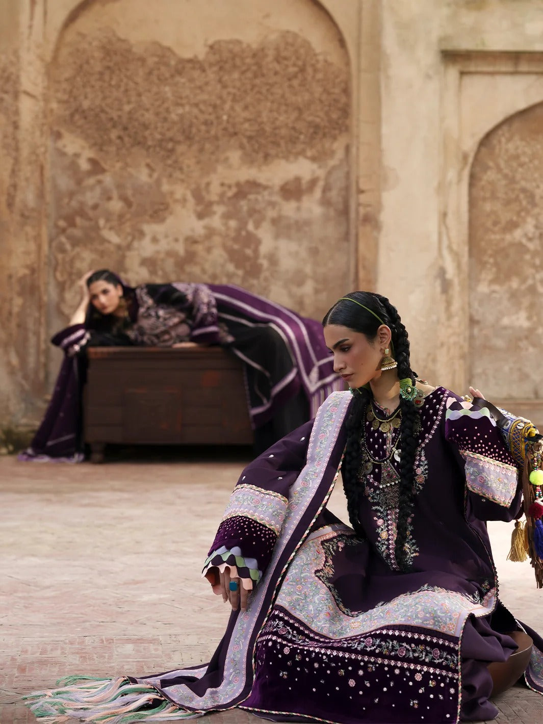 A woman in a traditional deep purple embroidered outfit kneels in a courtyard, while another person lies on a wooden bed in the background, both draped in matching purple fabric. The setting features textured beige stone walls and arches.