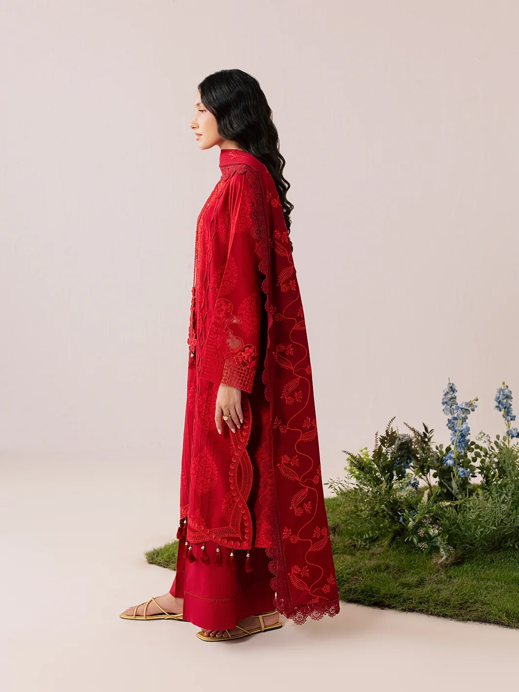 A woman in profile wearing a long red embroidered kurta with matching pants and a sheer red dupatta, standing on a beige studio set with a small patch of green grass and blue flowers at the lower right.