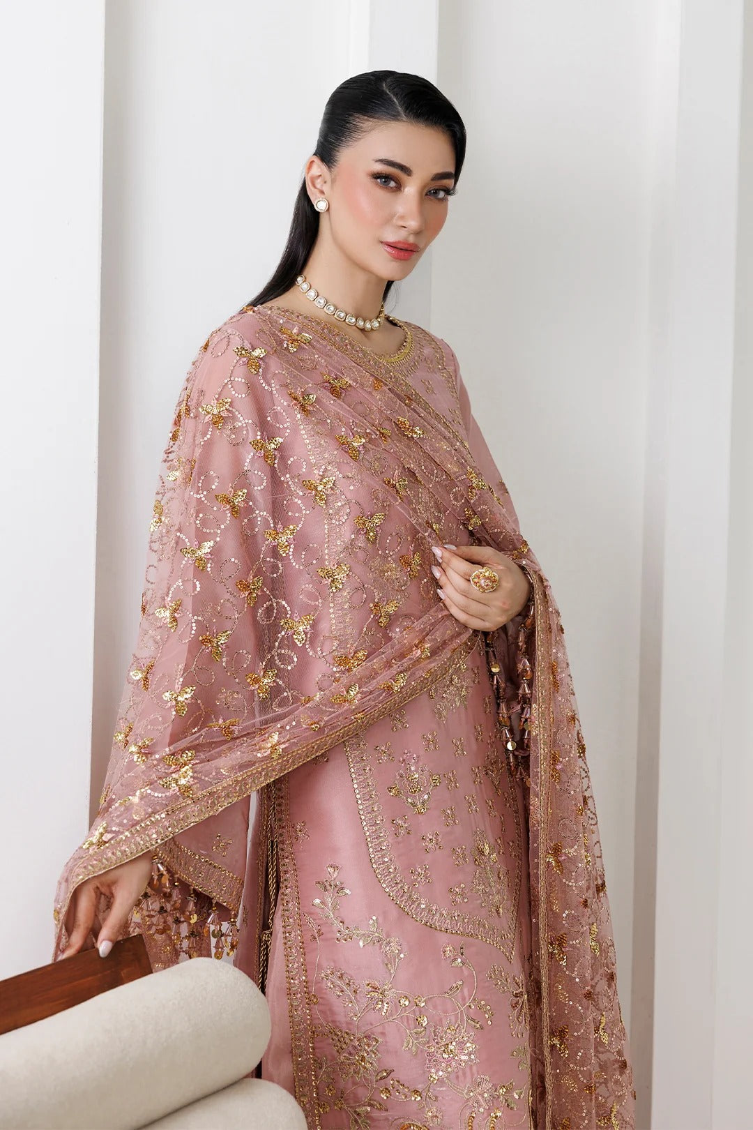 A woman models a dusty pink traditional outfit with intricate gold embroidery and a matching sheer dupatta draped over her shoulders, set against a clean white paneled background.