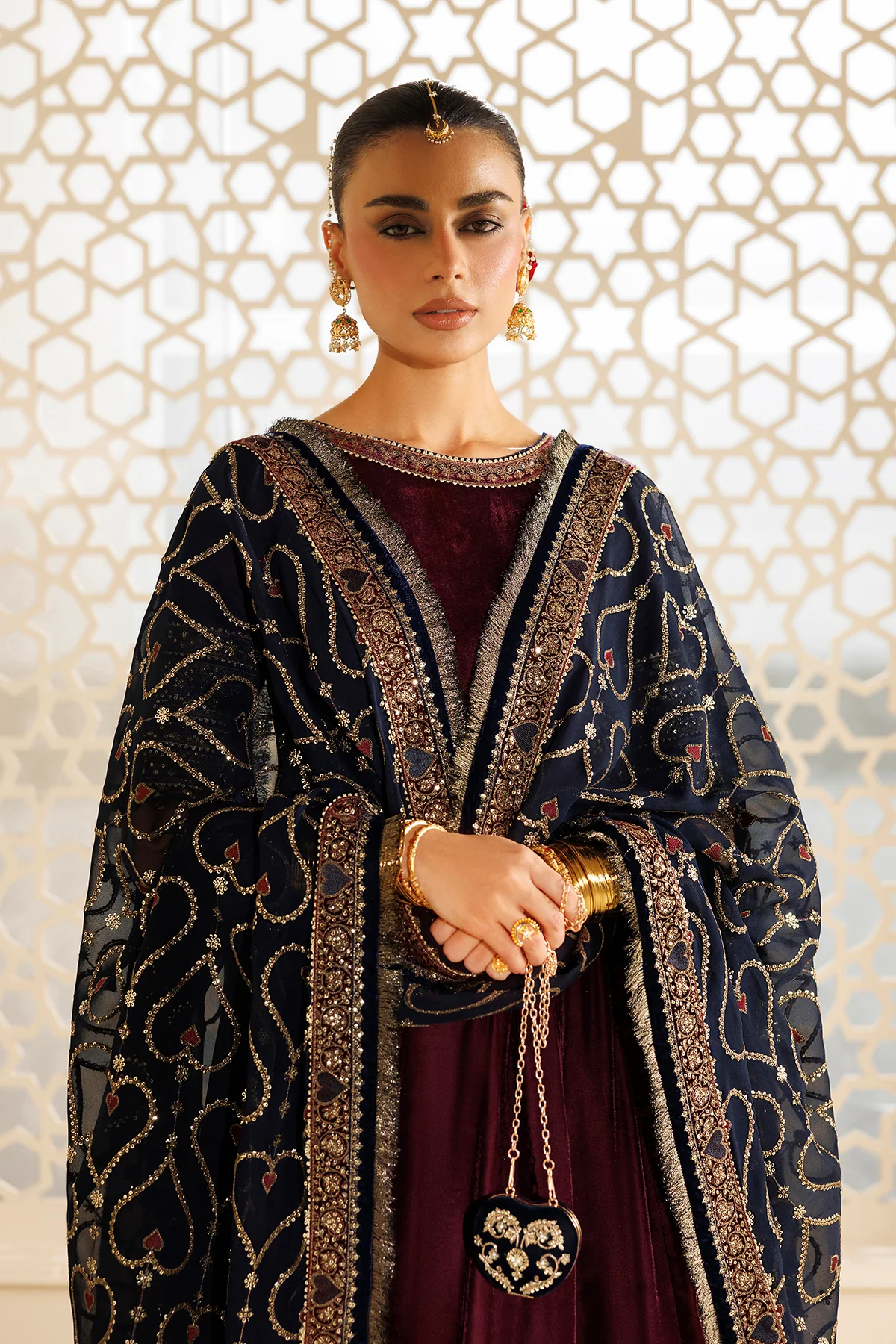 A woman models a traditional South Asian outfit featuring a navy blue heavily embellished dupatta with gold and silver embroidery, draped over a deep maroon velvet dress. She is accessorized with gold jewelry, including a maang tikka, earrings, and a small heart-shaped purse, set against a warm, patterned screen background.