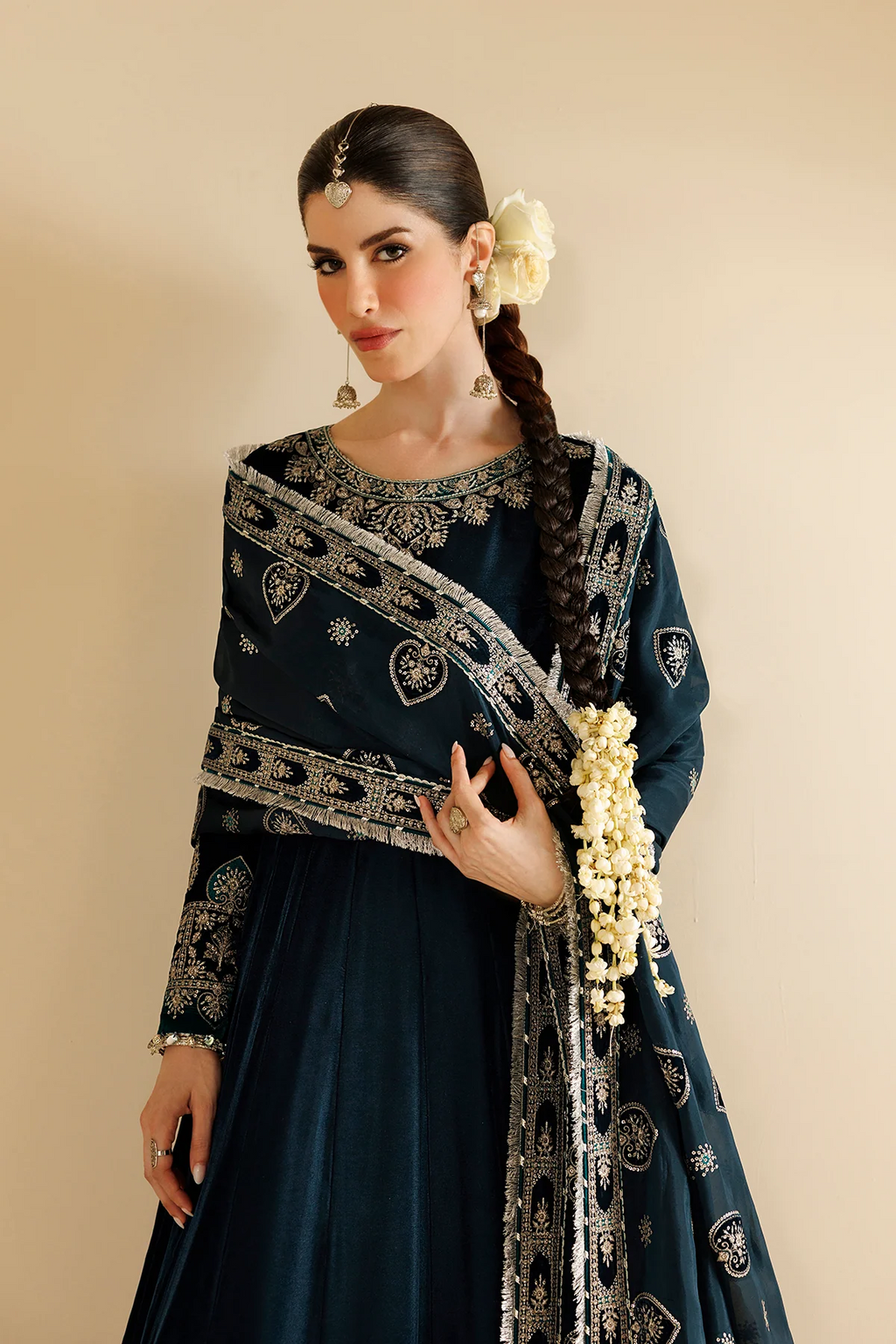 A woman models a dark teal blue traditional outfit with intricate silver embroidery and a matching dupatta, styled with a white flower in her hair and white jasmine garlands on her wrist, set against a solid beige background.