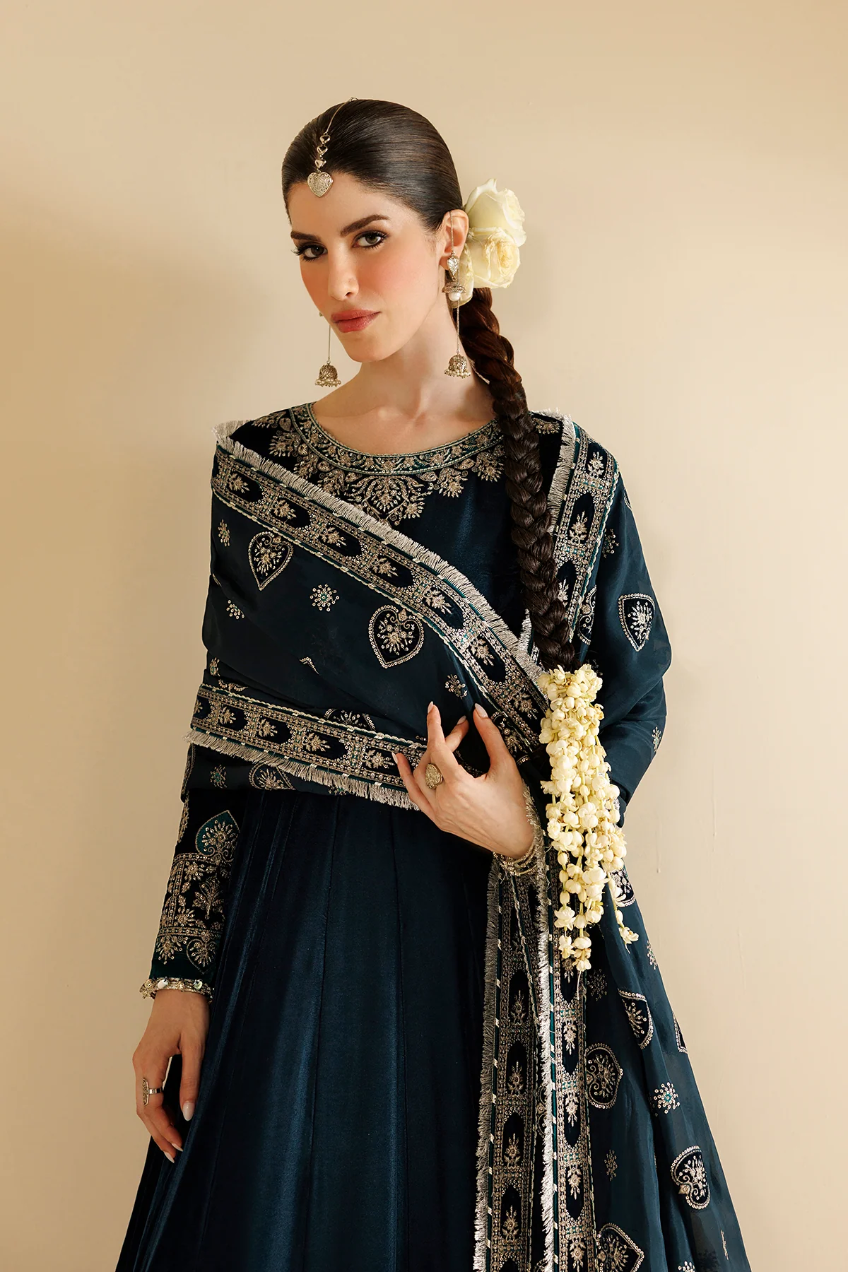 A woman models a dark teal blue traditional outfit with intricate silver embroidery and a matching dupatta, styled with a white flower in her hair and white jasmine garlands on her wrist, set against a solid beige background.