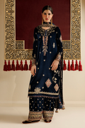 A woman models a dark navy blue traditional South Asian outfit with intricate gold and silver embroidery, paired with matching embroidered pants and a dupatta, set against an ornate gold and red background.