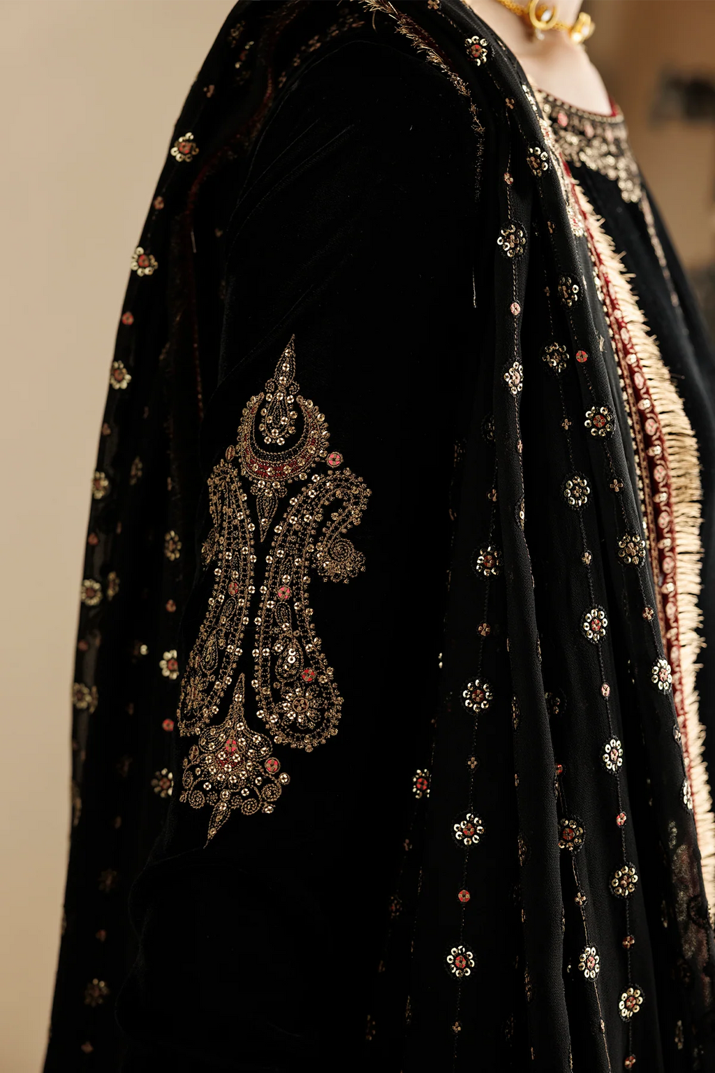 Close-up of a black embroidered traditional garment with ornate metallic and gemstone detailing, including a large decorative motif on the sleeve and scattered floral embroidery on the fabric.