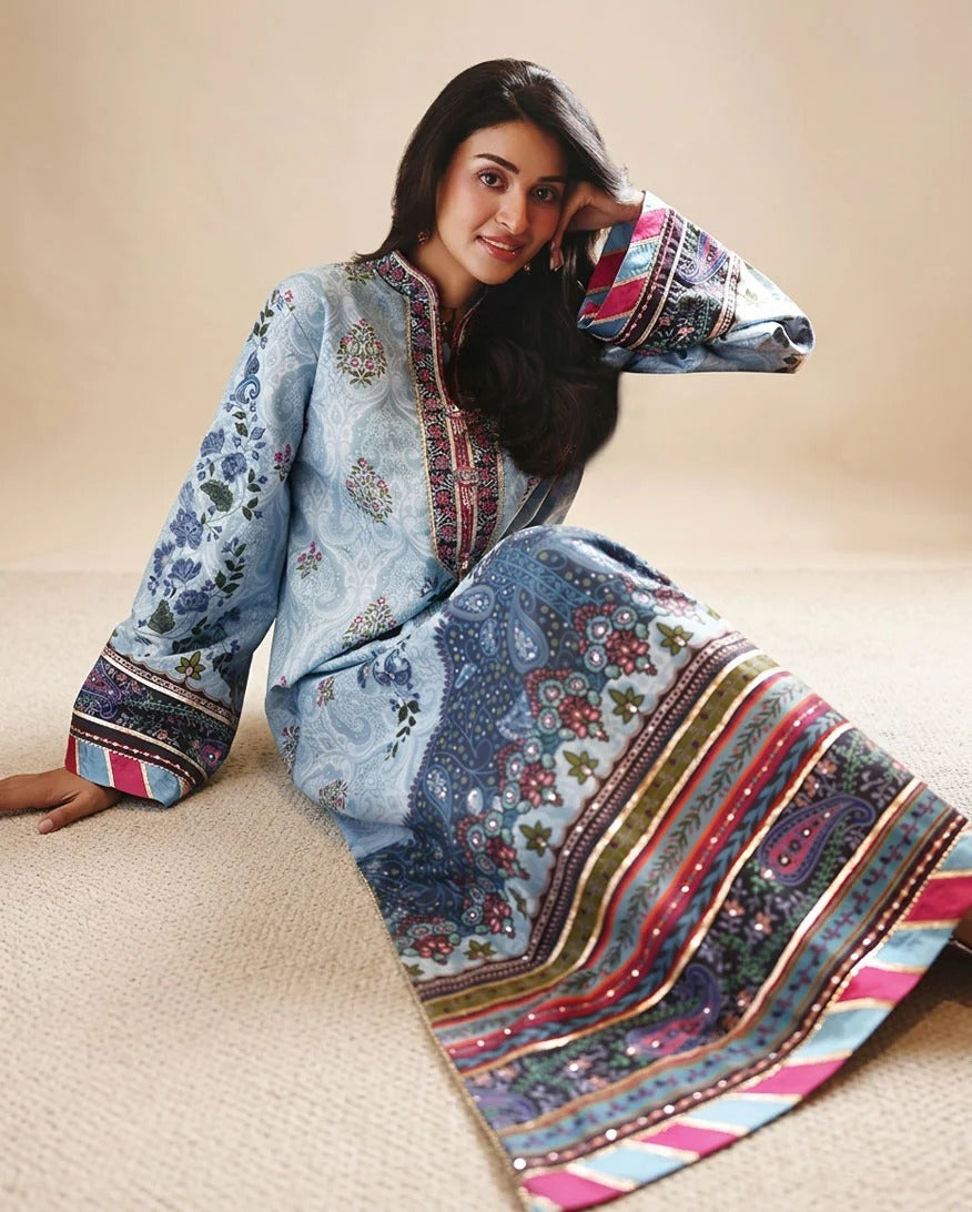 A woman wearing a light blue embroidered kurta with multicolor paisley and striped patterns, sitting on a beige carpet against a neutral beige background.