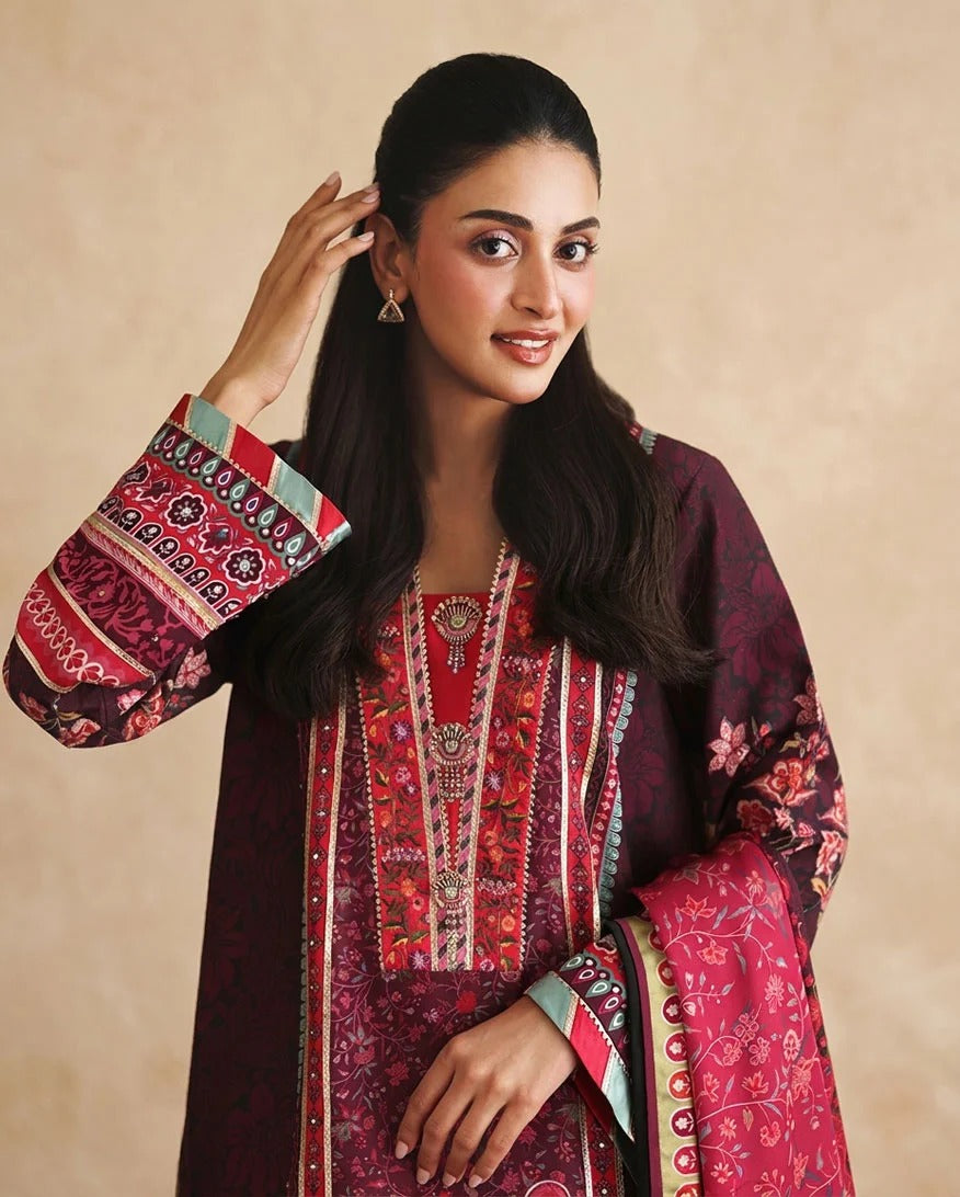 A woman modeling a traditional South Asian salwar kameez suit in deep red and maroon with intricate floral patterns, a matching dupatta, and patterned sleeve cuffs, set against a warm beige background.