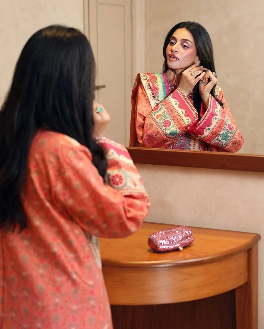 A woman with long black hair looks into a mirror while wearing a coral-pink patterned kurta. Her reflection shows her adjusting a colorful patterned necklace.
