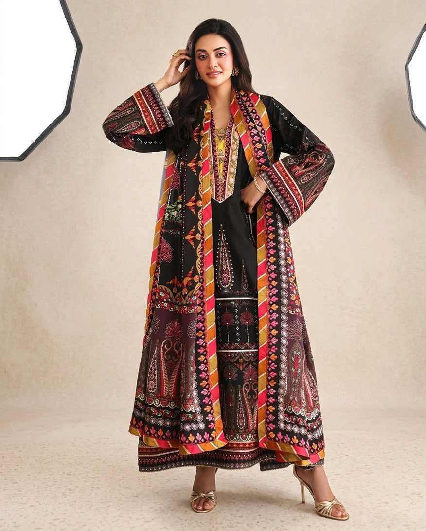 A woman modeling a traditional South Asian outfit consisting of a black kurta with intricate red, yellow, and gold patterns, paired with matching wide-leg trousers and a long open-front patterned jacket. She is standing against a neutral beige studio backdrop with hexagonal light panels visible on the sides.