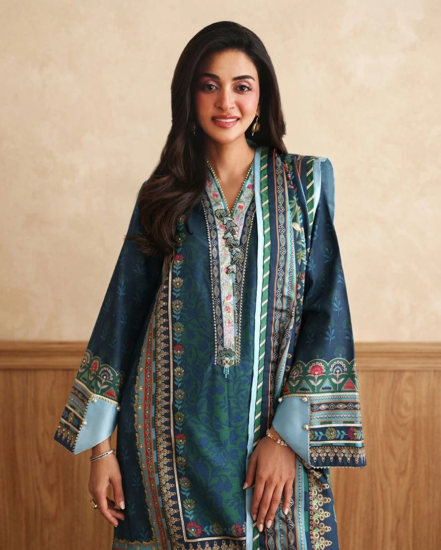 A woman modeling a traditional South Asian salwar kameez set in teal blue with intricate multicolor embroidery, featuring a matching embroidered dupatta, set against a beige textured wall and wooden paneling.