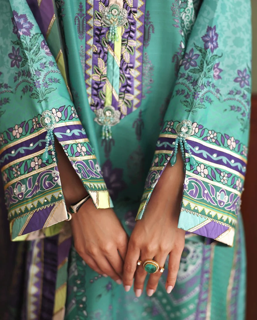 A close-up of a person wearing a richly patterned traditional outfit in shades of teal green, purple, and gold. The fabric features intricate floral and geometric prints, and the person is holding their hands together, showcasing a gold ring with a green stone.