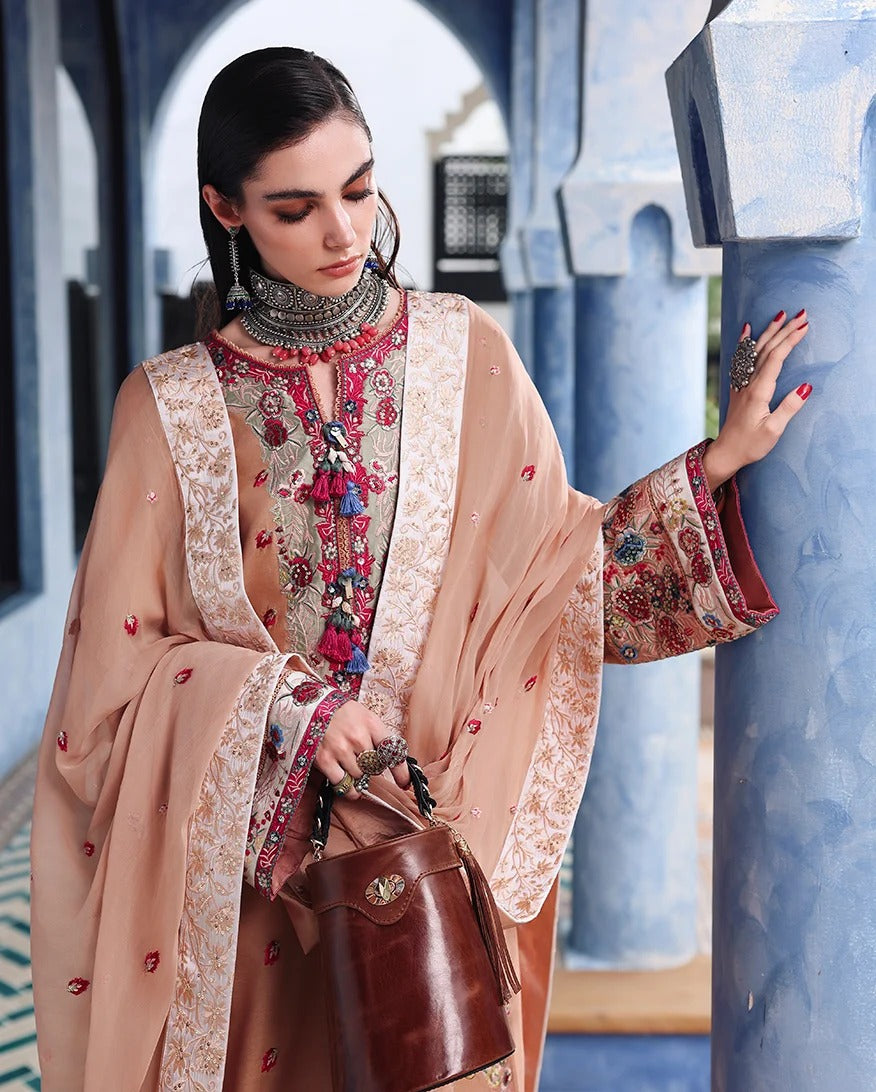 A woman models a traditional Moroccan-style outfit featuring a beige embroidered tunic with colorful floral patterns and a matching sheer beige shawl draped over her shoulders. She is posed in front of blue arches, holding a brown leather handbag.