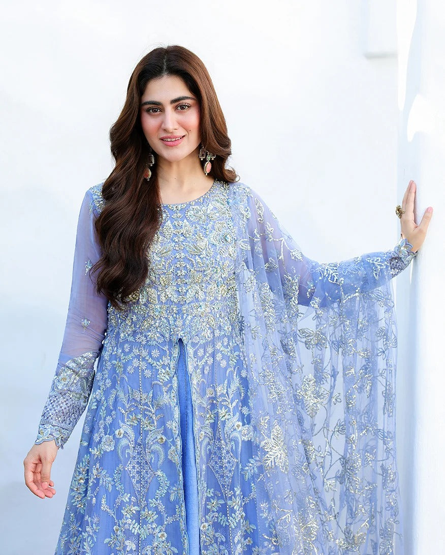 A woman wearing a light blue embroidered traditional outfit with sheer sleeves and a matching draped overlay, posed against a bright white wall.